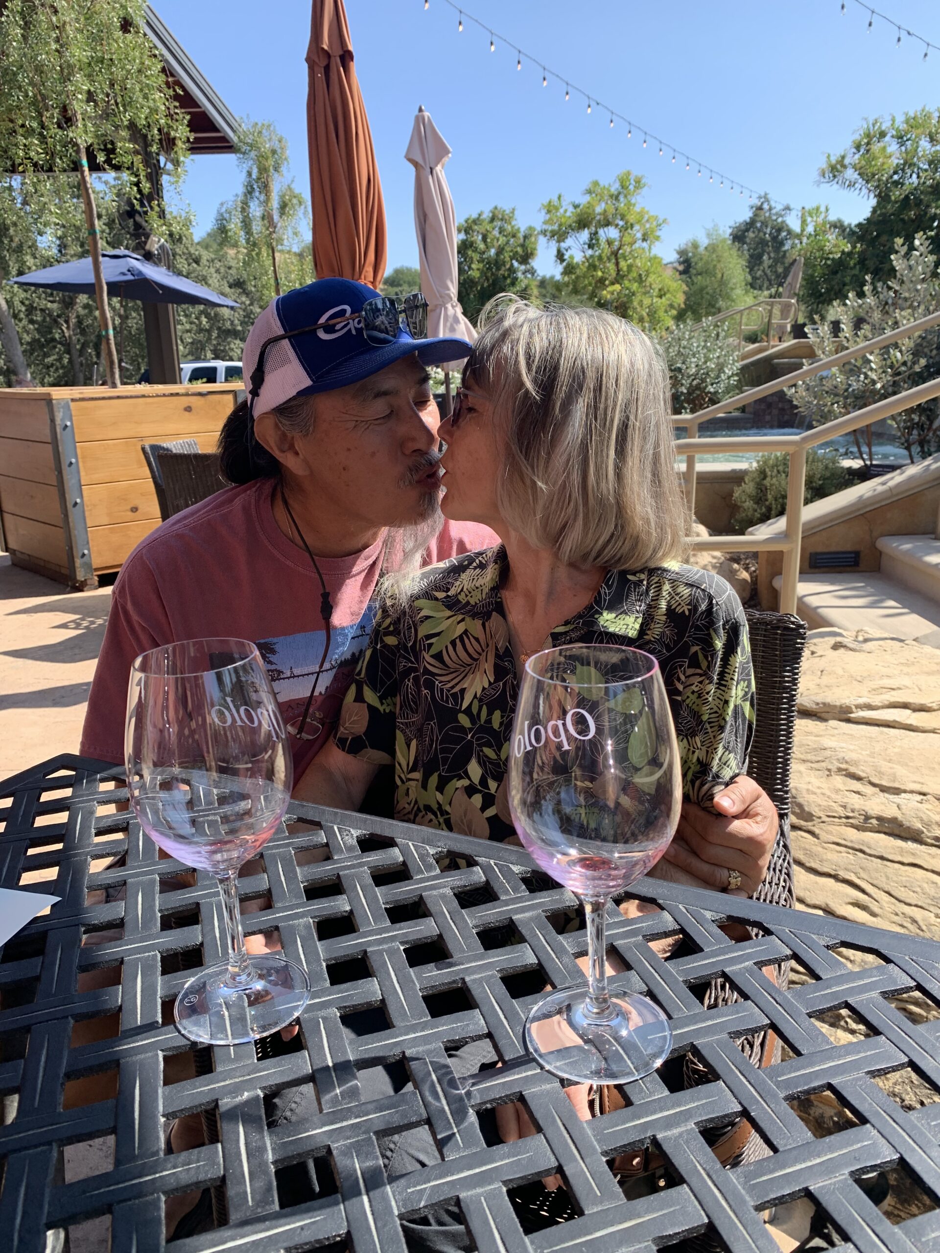 Two people about to kiss at an outdoor table with two empty wine glasses on it